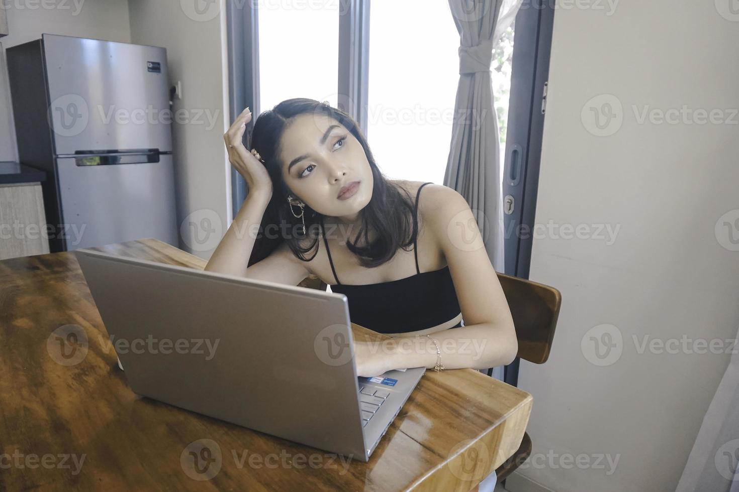 Young Asian woman spends her time at home sitting in the dining room frustrated and tired working on her laptop. photo