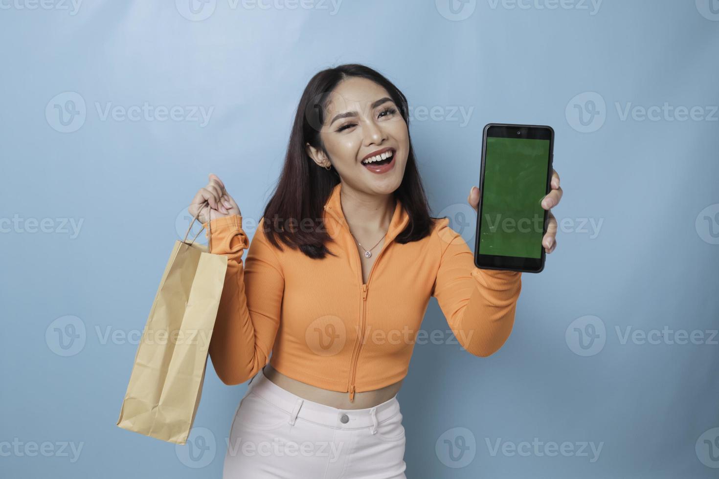 Portrait Asian happy beautiful young woman standing excited holding an online shopping bag and showing her smartphone, studio shot isolated on blue background photo