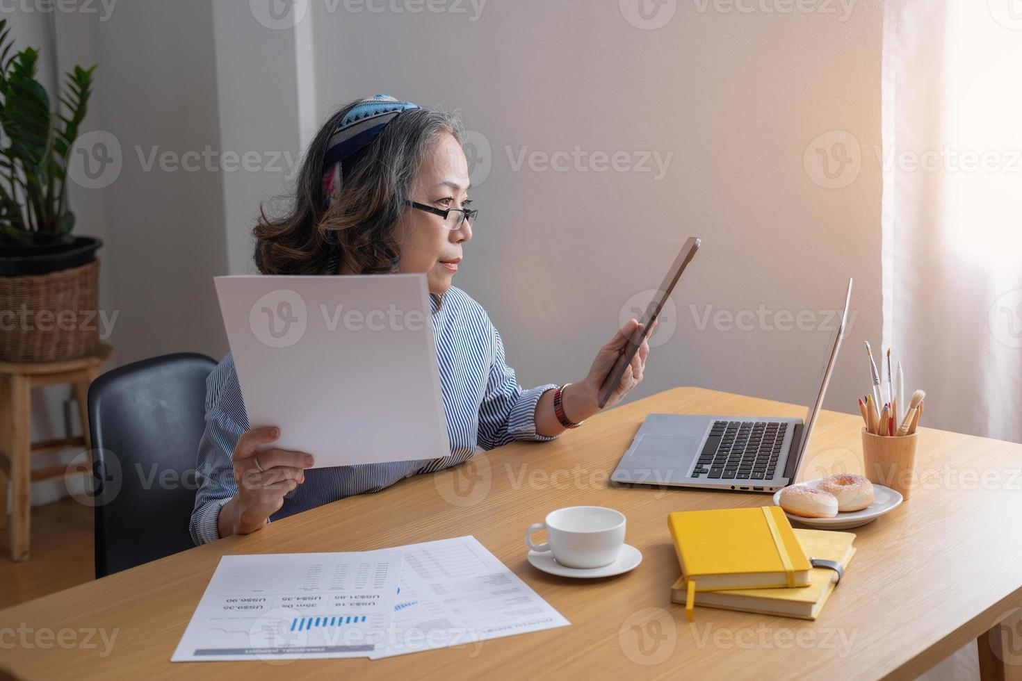 Portrait of an elderly woman working inside the house using laptops and tablets to work and check important documents for customers. photo