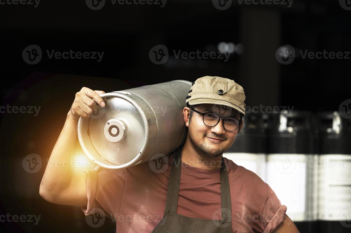 Young man in leather apron holding beer keg at modern brewery, craft brewery worker photo