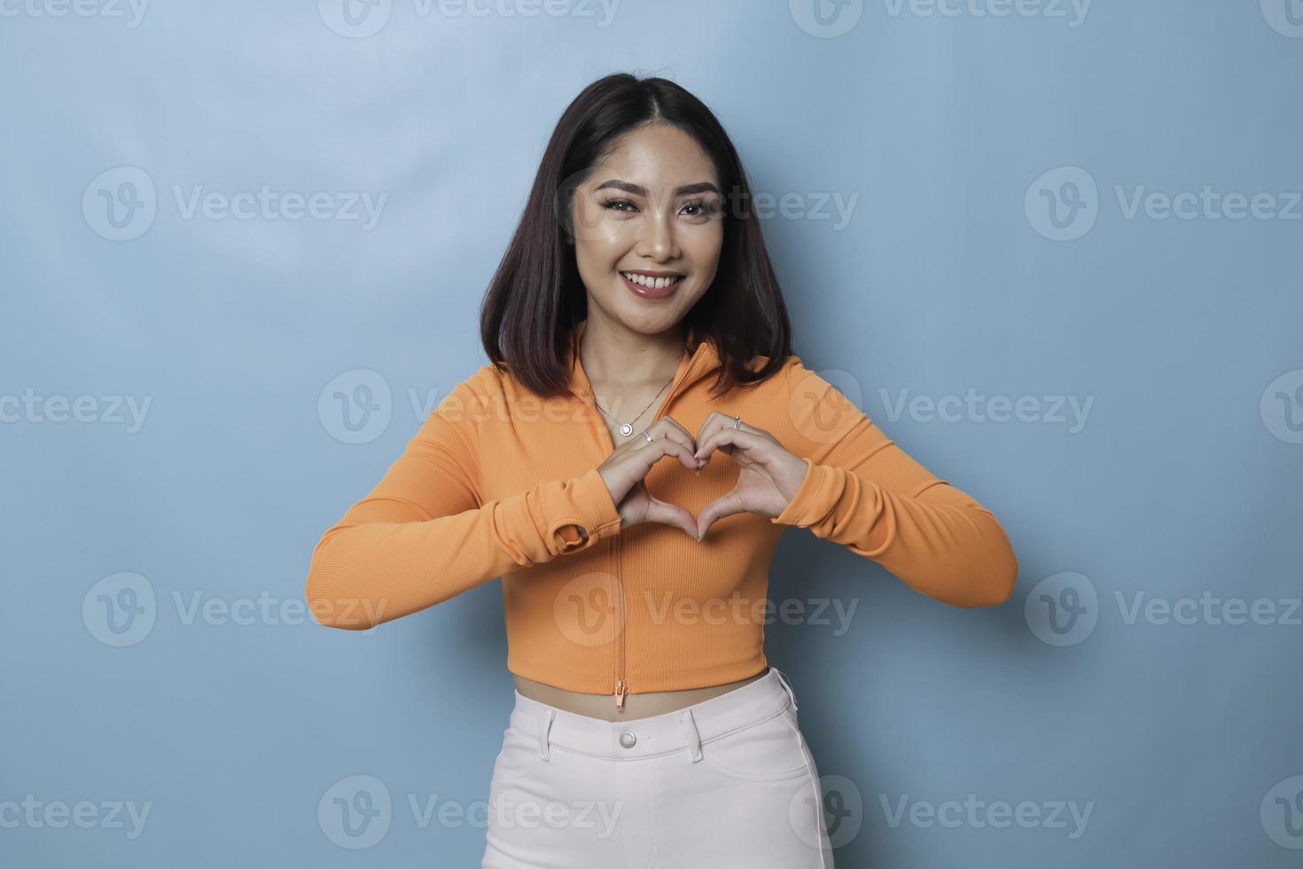 Attractive young Asian woman feels happy and romantic shapes heart gesture expresses tender feelings wears casual orange top against blue background. People affection and care concept photo