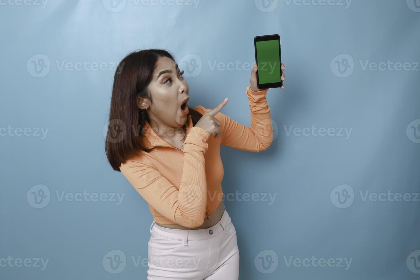 A surprised young woman pointing at copy space on her smartphone in her hand, isolated on blue background photo