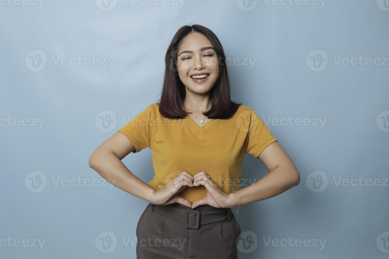 Attractive young Asian woman feels happy and romantic shapes heart gesture expresses tender feelings wears casual yellow t-shirt against blue background. People affection and care concept photo