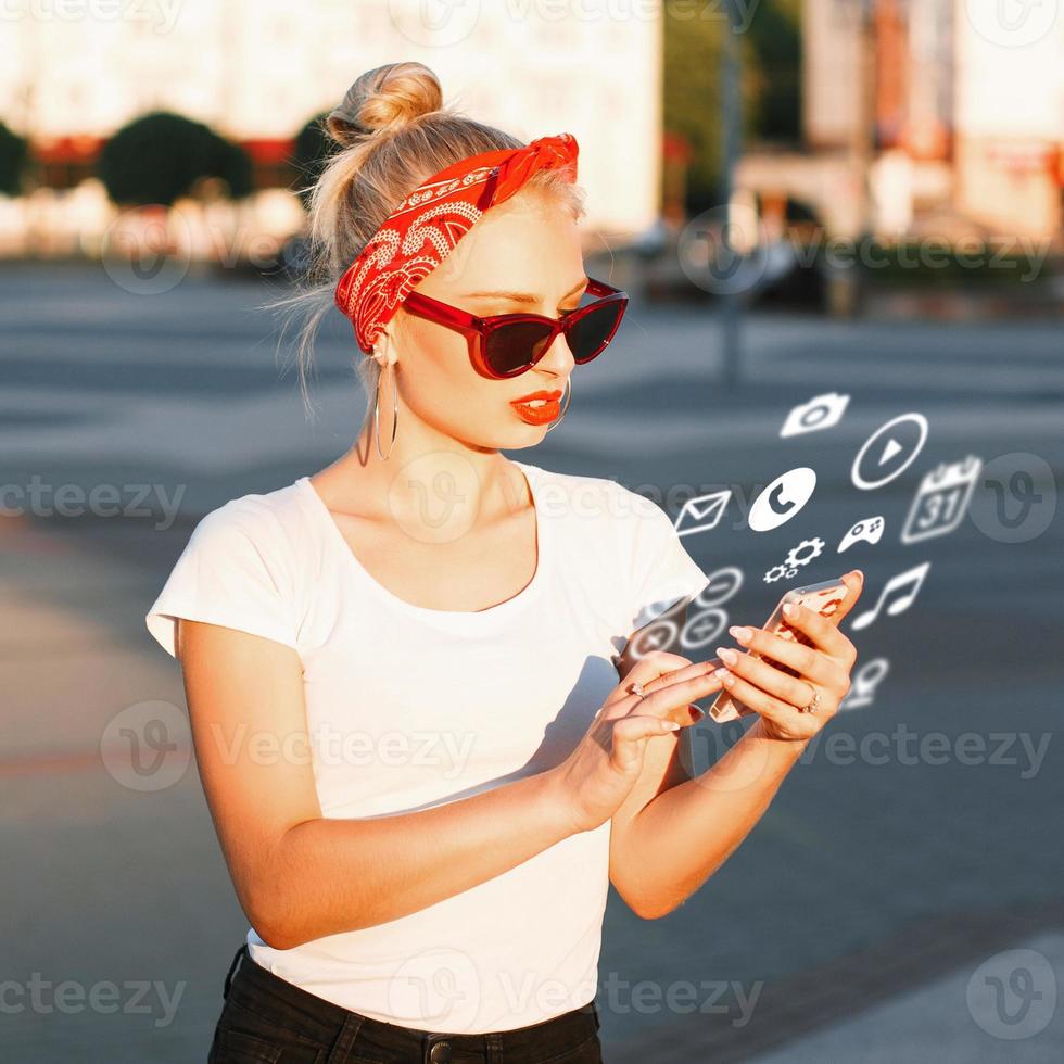 hermosa chica hipster con gafas de sol sosteniendo un teléfono con iconos voladores. foto