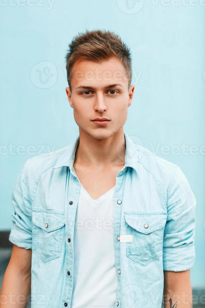 retrato de primer plano de un joven con estilo en una camisa de mezclilla cerca de una pared azul. foto