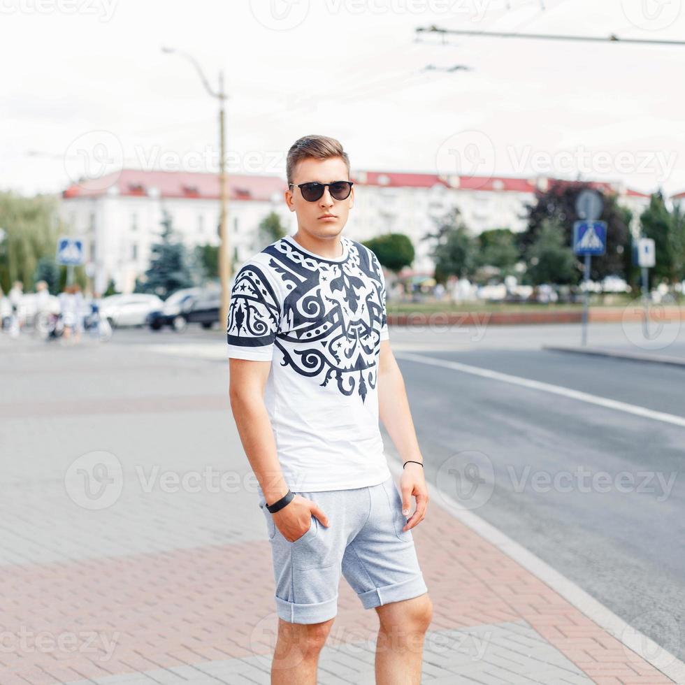 Handsome young man in T-shirt and shorts posing on the background of the city. photo
