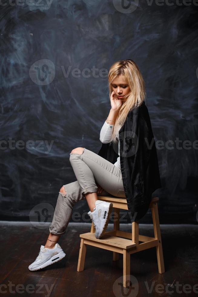 Pretty woman with blonde hair in a stylish black shirt, white sneakers and torn jeans sitting on a wooden chair on a dark background with chalk stains photo