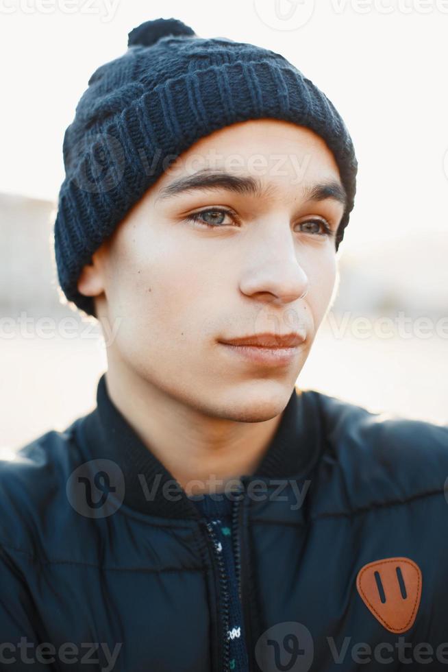 Close-up portrait of a young man in a warm knitted hat and winter jacket on a background of a sunset photo
