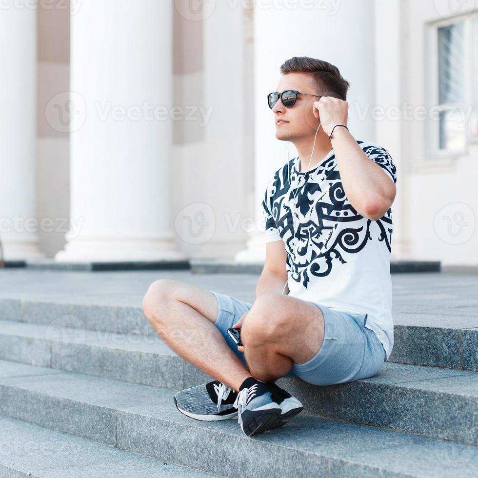 Young stylish man with sunglasses sitting on the stairs and listening to music. photo