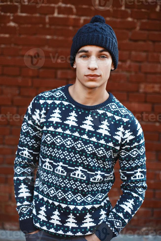 Young stylish hipster in a knitting sweaters with patterns and a hat on a background of red brick wall photo