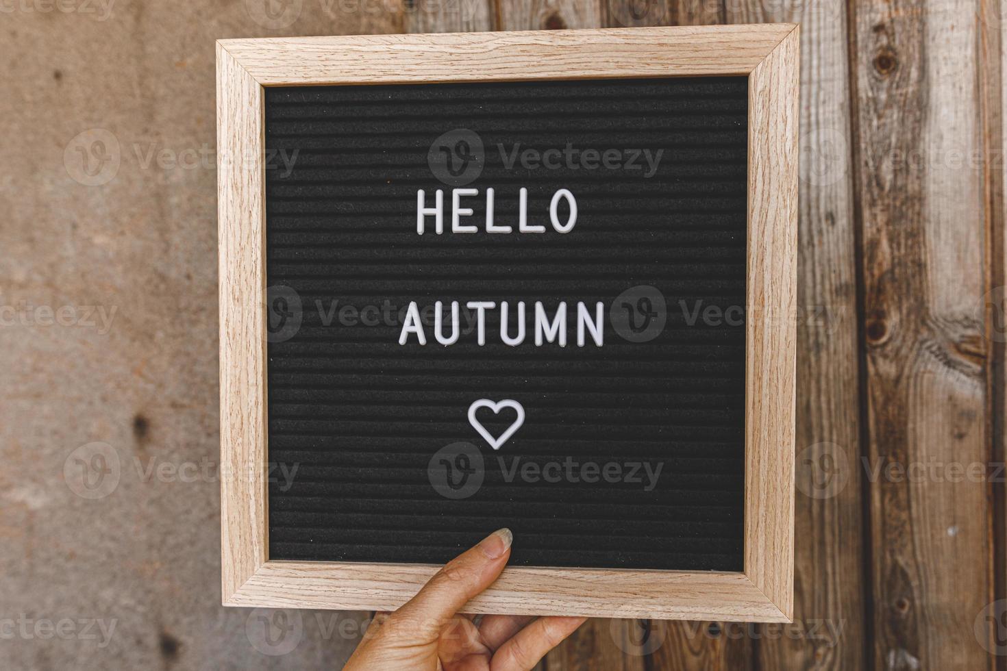 Autumnal Background. Woman hand holding black letter board with text phrase Hello Autumn on wooden palnks backdrop. Thanksgiving banner. Hygge mood, cold weather concept photo