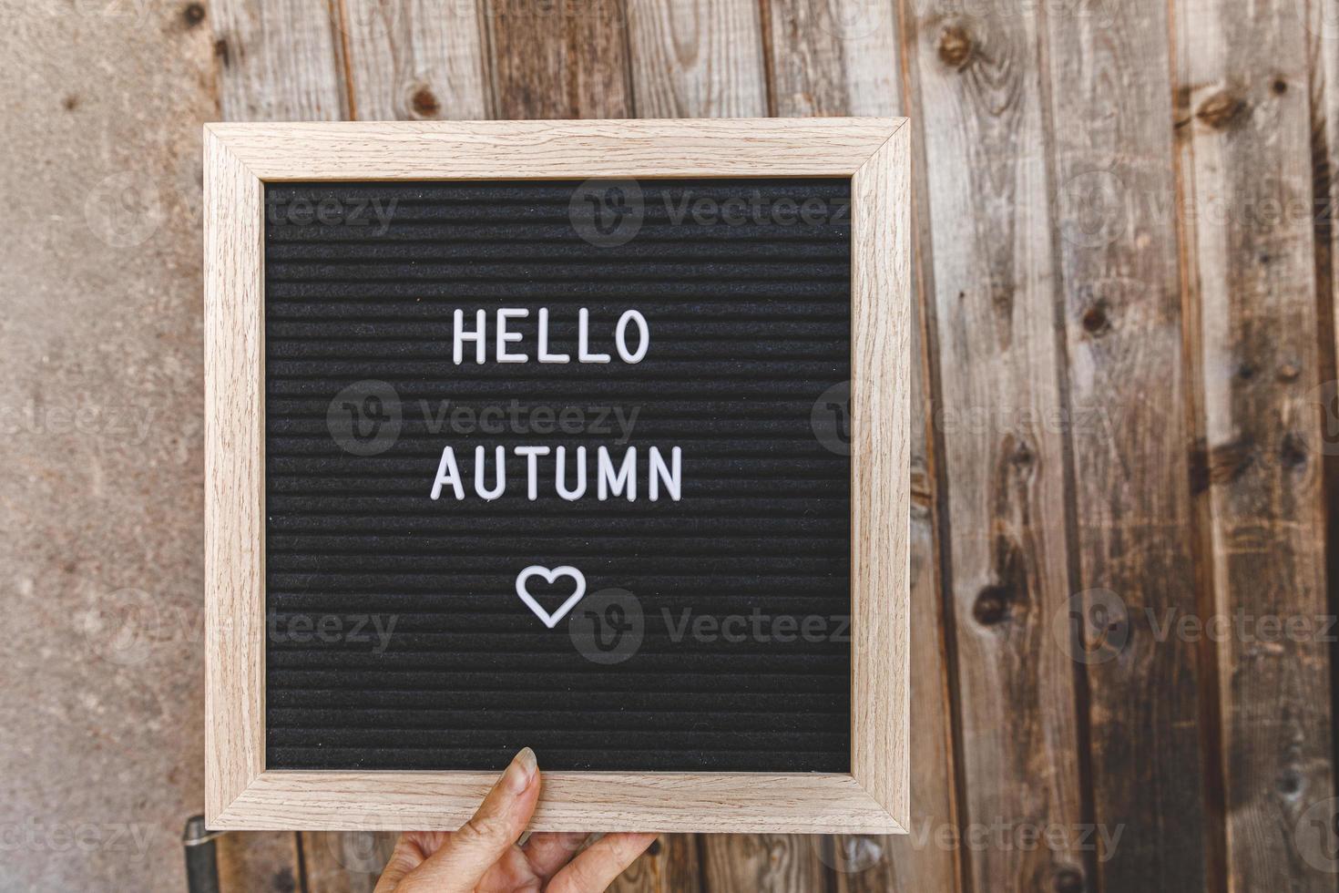 Autumnal Background. Woman hand holding black letter board with text phrase Hello Autumn on wooden palnks backdrop. Thanksgiving banner. Hygge mood, cold weather concept photo