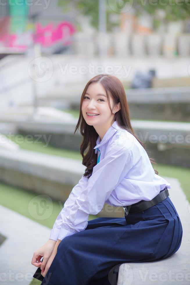 bella estudiante asiática de secundaria con uniforme escolar con frenos en los dientes sentada y sonríe con confianza mientras mira a la cámara felizmente con el edificio en el fondo. foto