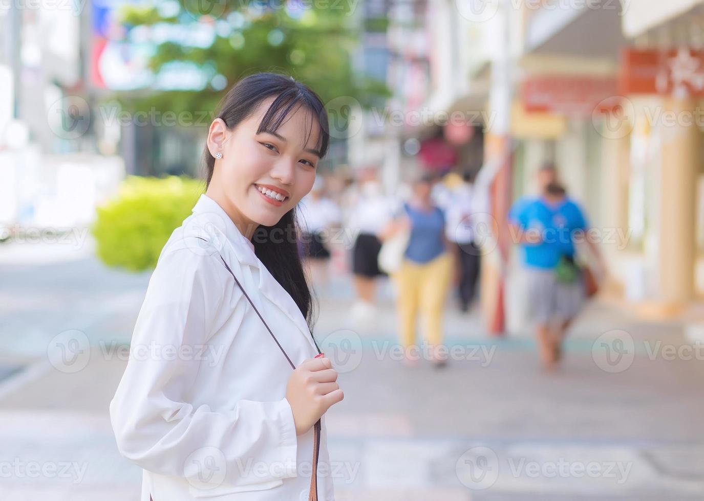 Confident young business Asian working female who wears a white shirt and shoulder bag smiles happily while she is walking to work at office in the city. photo