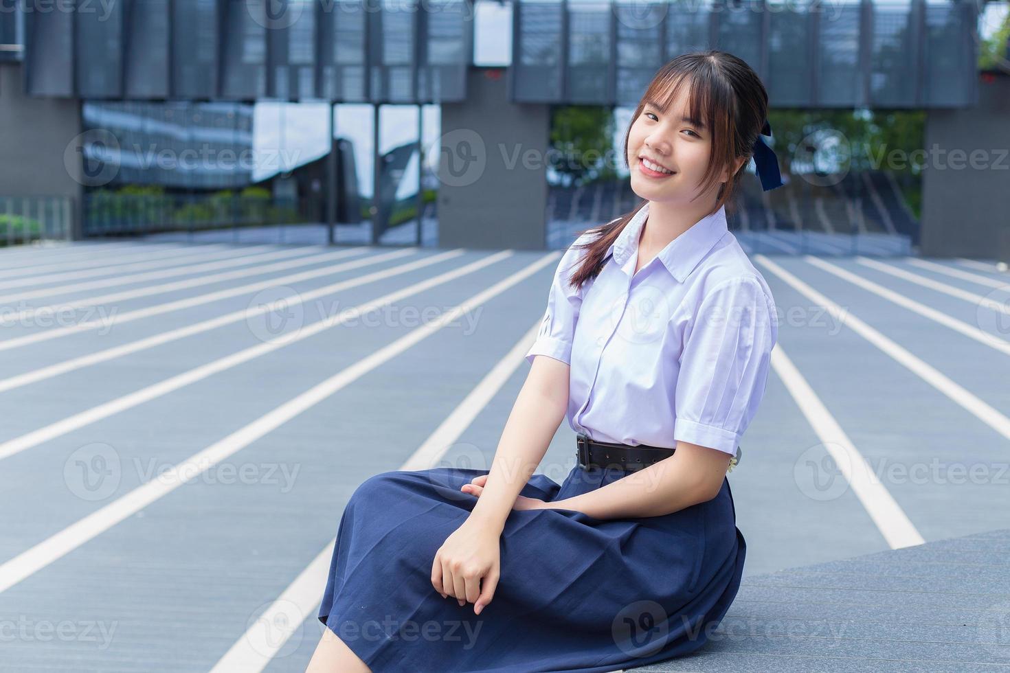 hermosa estudiante asiática de secundaria con uniforme escolar con sonrisas confiadas mientras mira la cámara felizmente con el edificio en el fondo. foto