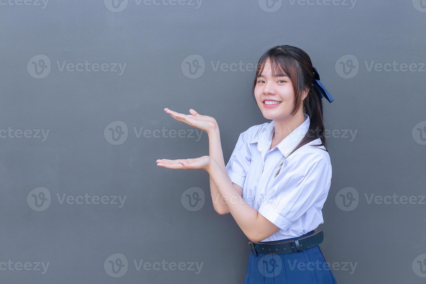 Cute Asian high school student girl in the school uniform with smiles confidently while she looks at the camera to present something happily with grey in the background. photo