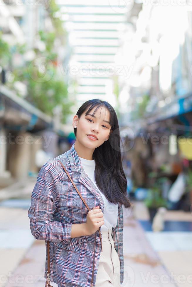 Confident professional young Asian business woman who wears a brown striped blazer and shoulder bag smiles happily and looks at the camera as she commute to work through the old town. photo