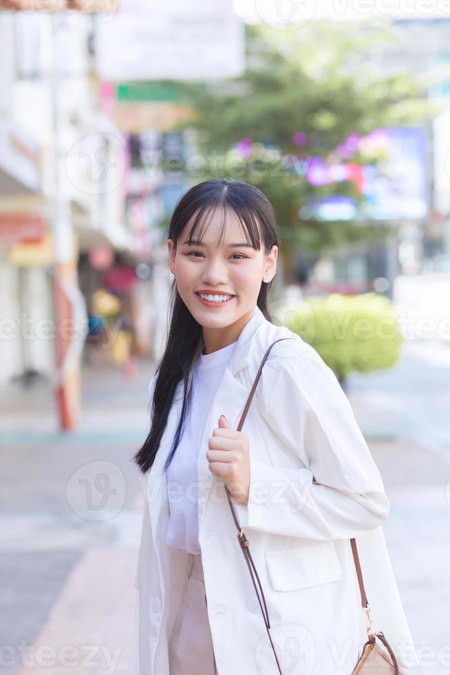 Confident young Asian female who wears a white shirt and shoulder bag smiles happily while she is walking to work at office in the city. photo
