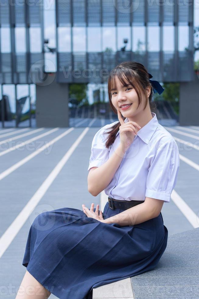 Beautiful Asian high school student girl in the school uniform with smiles confidently while she sits looks at the camera happily with the building in the background. photo