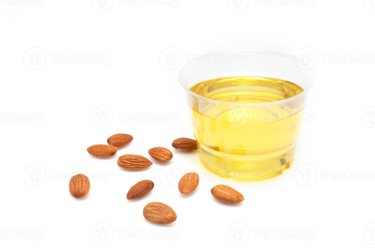 Yellow almond oil in a clear bowl with a wooden spoon next to it and several almonds on a white background. photo