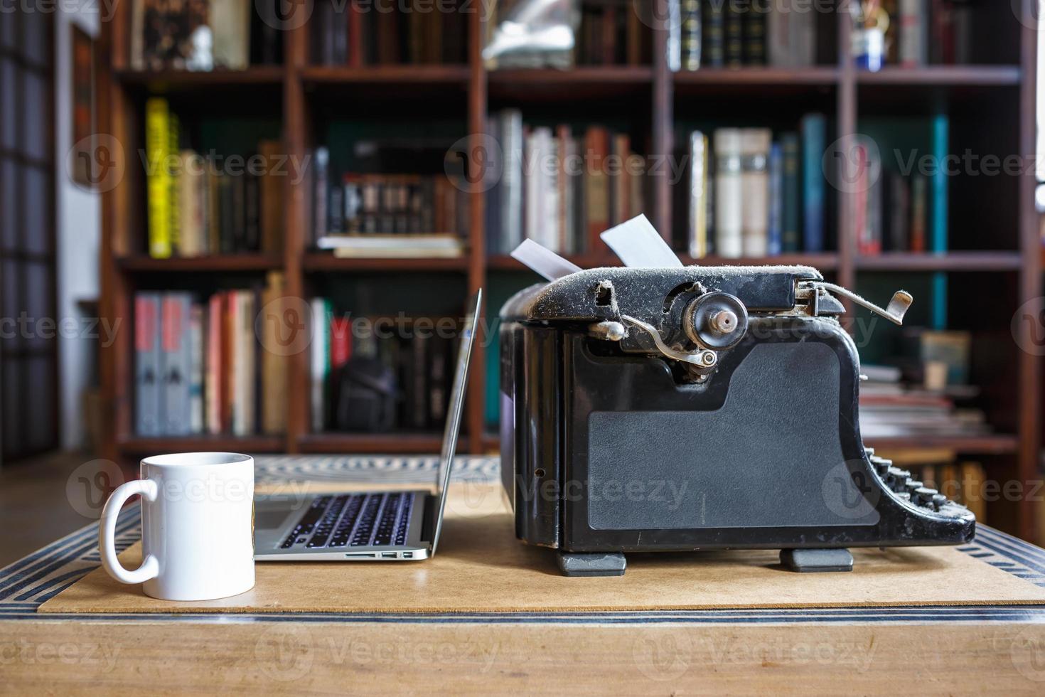 old vintage dust-covered typewriter with sheet of white paper near modern notebook and cup of coffee on bookcase background. modern technology and vintage appliances photo
