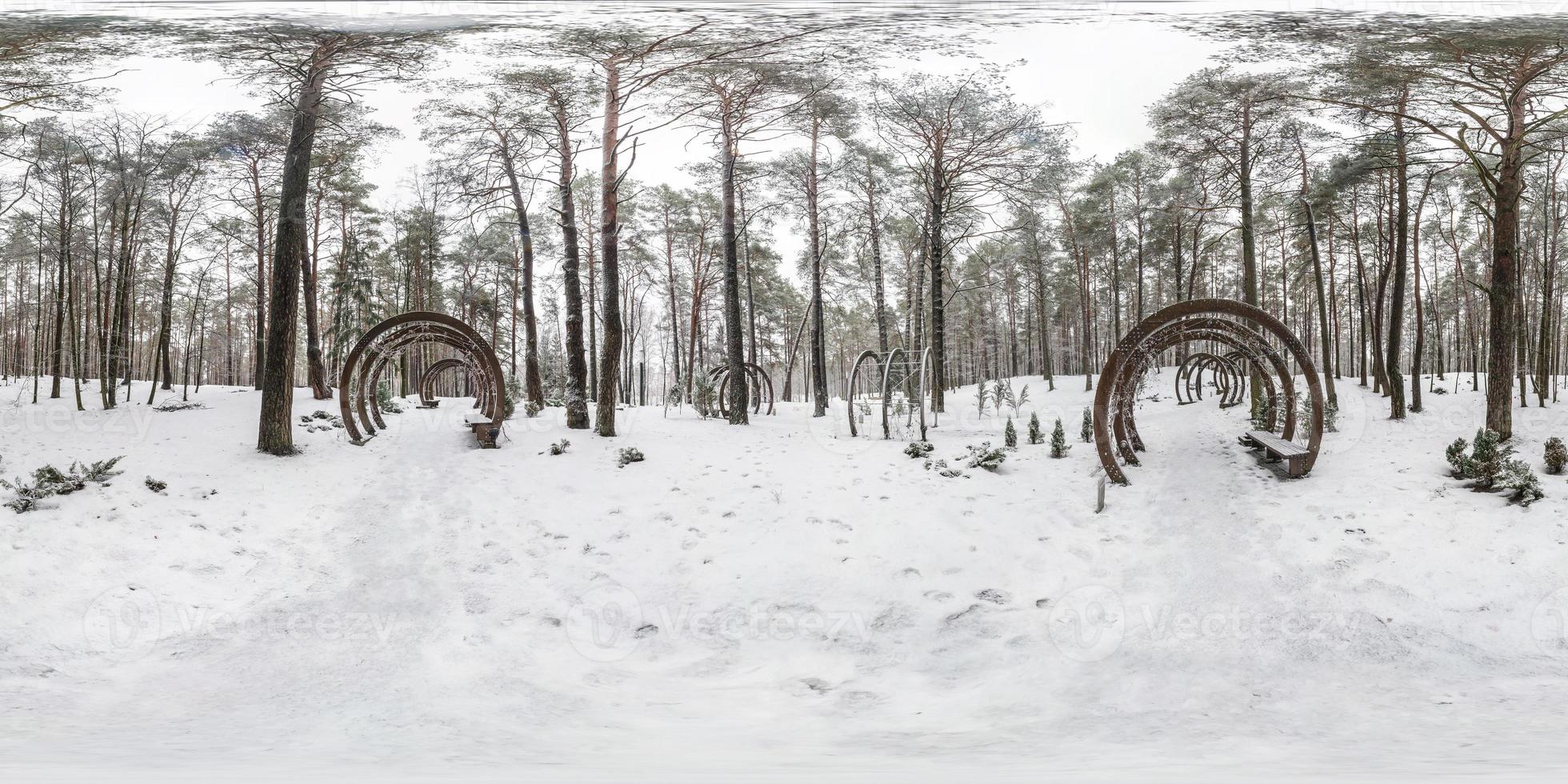 Winter full spherical seamless panorama 360 degrees angle view on road in a snowy park with gray pale sky near arches and benches in equirectangular projection. VR AR content photo