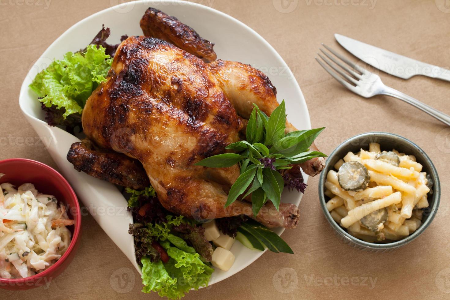 Chicken rotisserie with pasta salads on the sides, knife and fork, closeup on a ceramic plate on the table. Horizontal top view from above photo