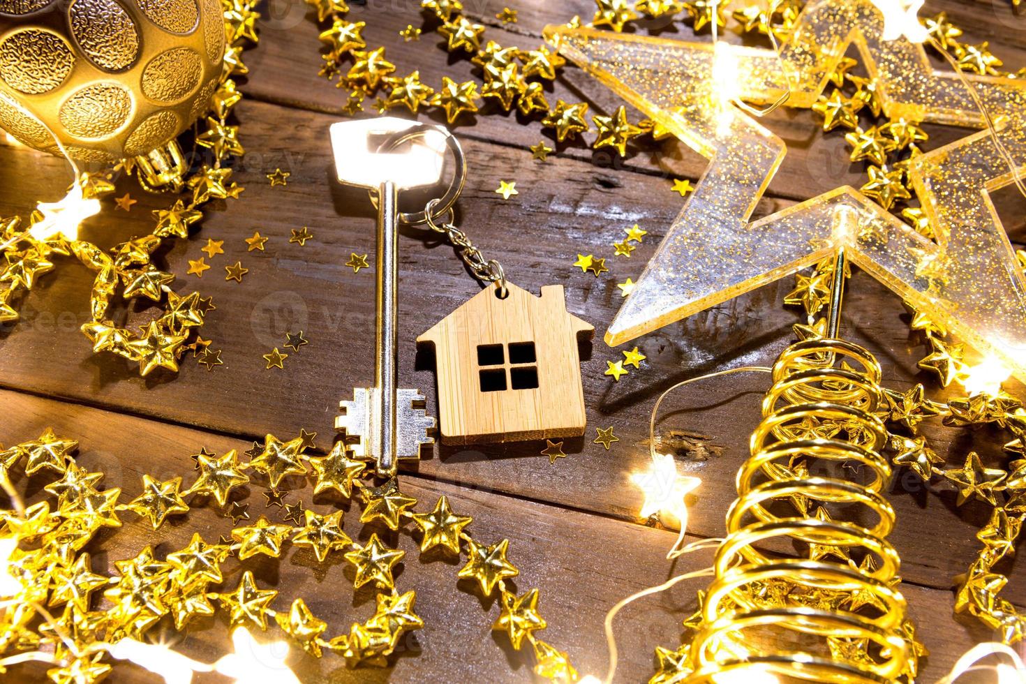 House key with keychain cottage on a festive background with sequins, stars, lights of garlands. Happy New Year-wooden letters, greetings, greeting card. Purchase, construction, relocation, mortgage photo