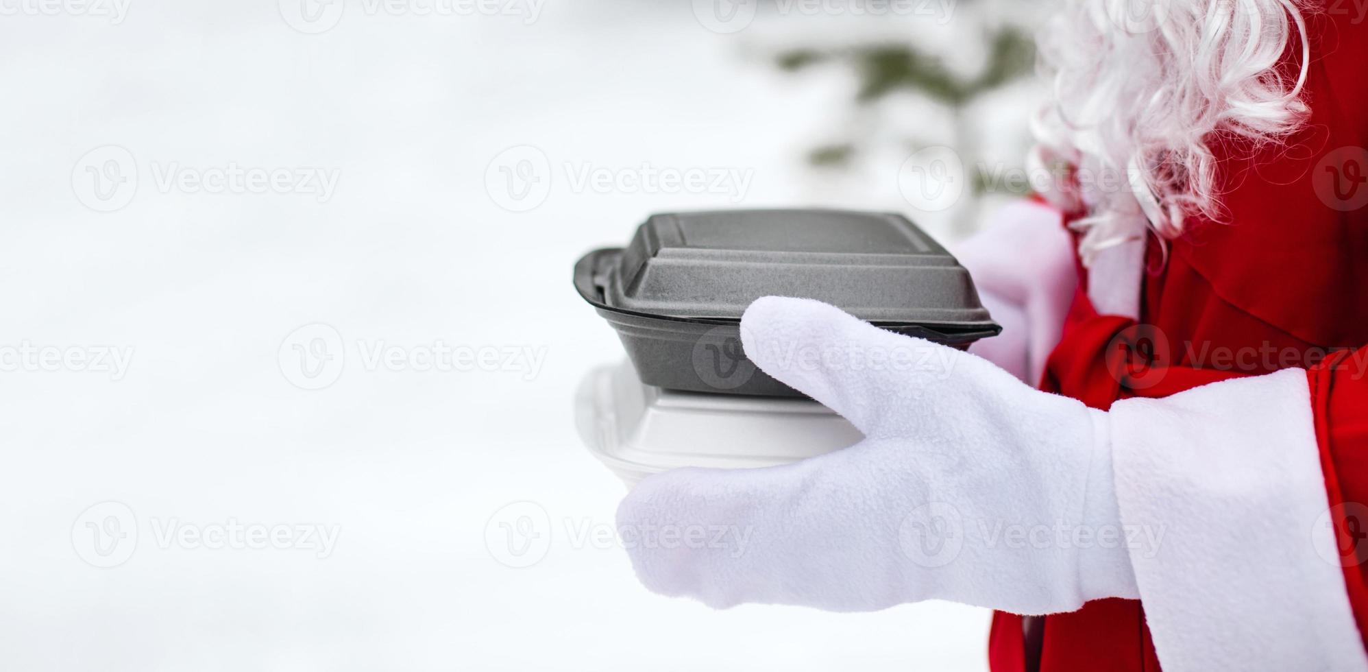 Food delivery service containers in hands of Santa Claus outdoor in snow. Christmas eve promotion. Ready-made hot order, disposable plastic box. New year holidays catering. Copy space, mock up photo