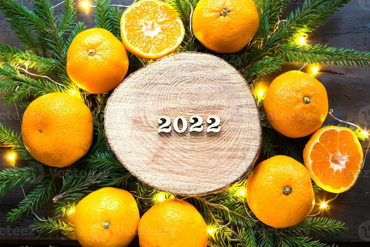 New year's holiday background on a round cut of a tree surrounded by tangerines, live fir branches and golden lights garlands, with wooden numbers date 2022. Citrus aroma, Christmas. Space for text. photo