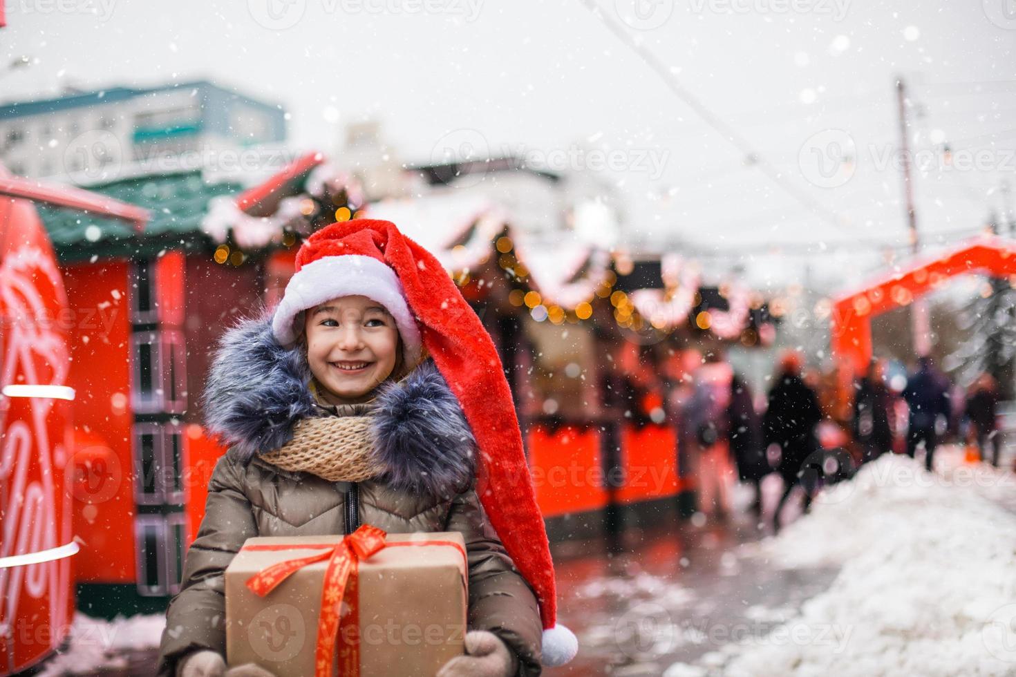 Portrait of joyful girl in Santa hat with gift box for Christmas on city street in winter with snow on festive market with decorations and fairy lights. Warm clothes, knitted scarf and fur. New year photo