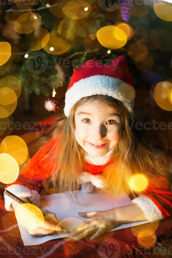 Little girl in a Santa hat and red dress under Christmas tree is dreaming, waiting for the holiday, lying on a plaid blanket. A letter on piece of paper, gifts. New year, Christmas. defocus lights photo