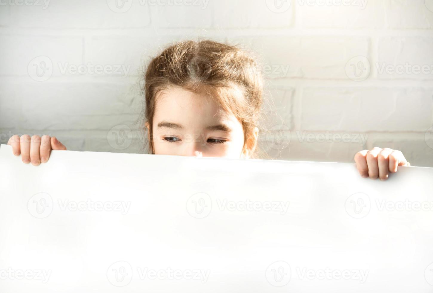 A sad girl holds a white sheet of paper - mock-up for advertising, slogan, inscription. Copy  space is in children's hands, the child is upset and hid. photo