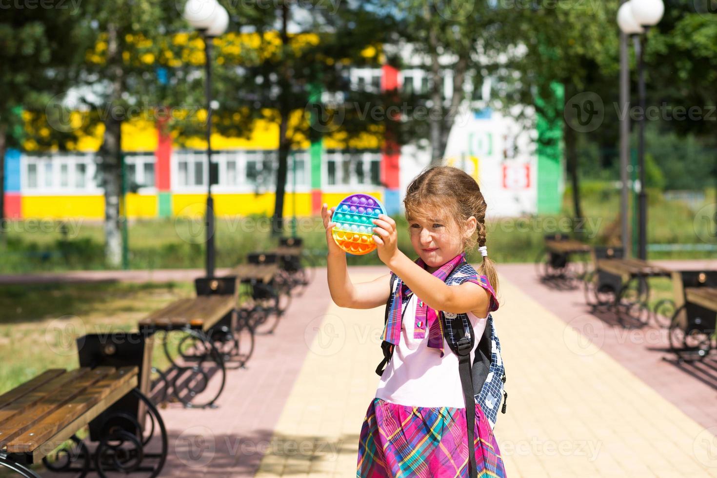 Little girl with a backpack and in a school uniform in the school yard plays pop it toy. Back to school, September 1. The pupil relaxes after lessons. Primary education, elementary class for student photo