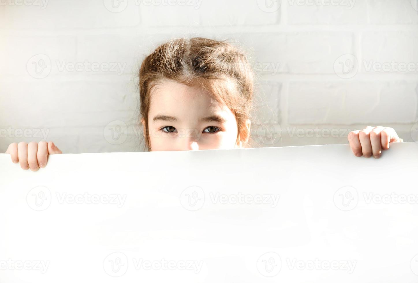 A sad girl holds a white sheet of paper - mock-up for advertising, slogan, inscription. Copy  space is in children's hands, the child is upset and hid. photo