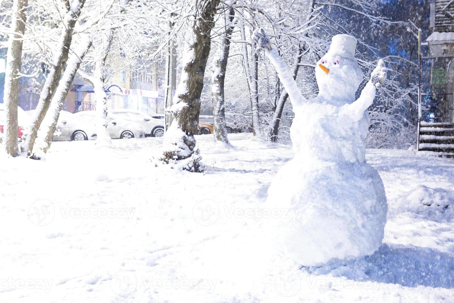 Big snowman in the yard after a snowfall in winter. Nose-carrot, bucket on the head, gloves on the hands, eyes. Cars in the snow along the road. Outdoor activities. Copy space photo