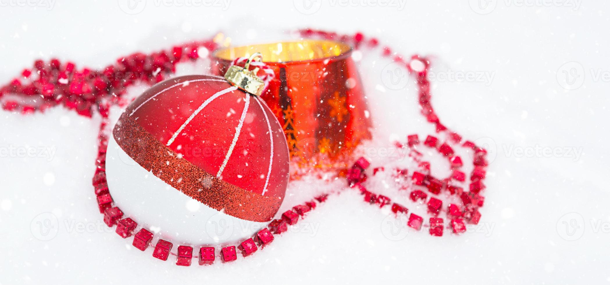Red Christmas ball on natural white snow with square beads and burning candle in a glass candlestick. Christmas, New year outdoor. Snowfall, festive mood of fairy tales and magic, street decoration. photo