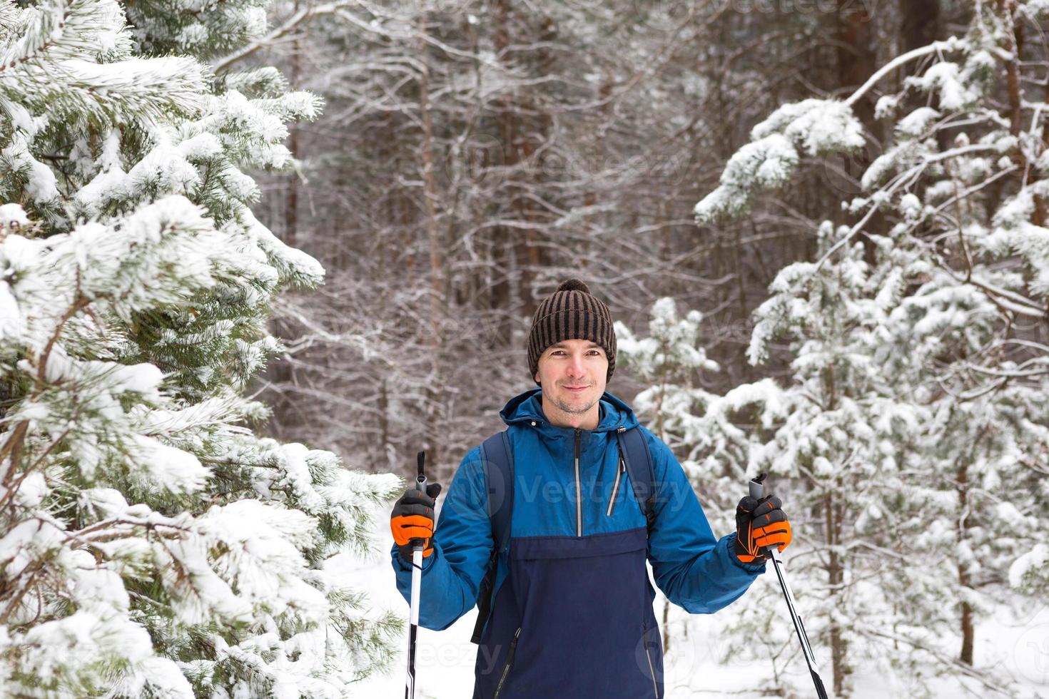 Skier with a backpack and hat with pompom with ski poles in his hands on background of a snowy forest. Cross-country skiing in winter forest, outdoor sports, healthy lifestyle, winter sports tourism. photo