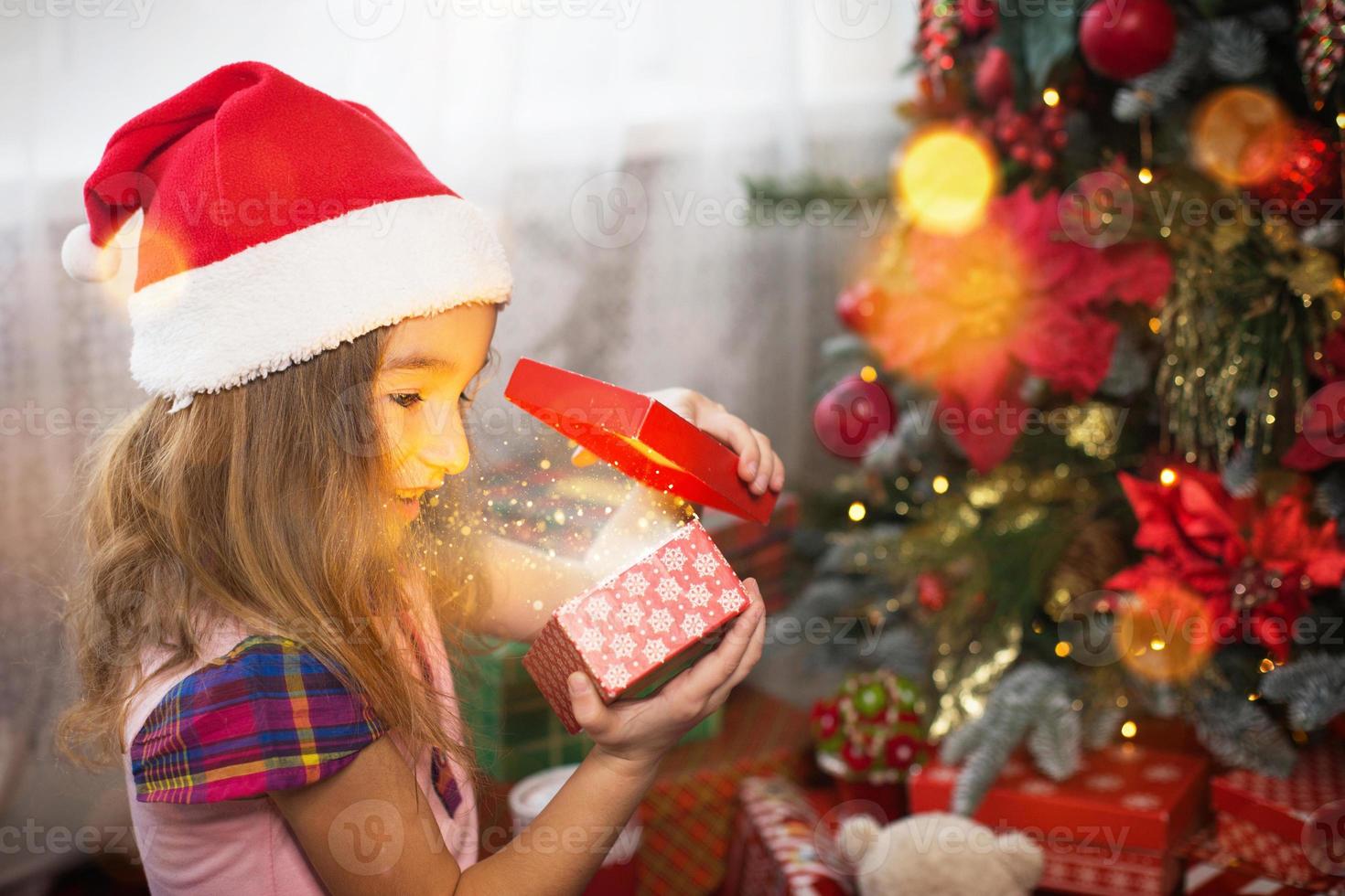 Little girl in Santa hat opens a red box with a gift and a Golden magic light near the Christmas tree. Holiday decor, poinsettias on fir trees, New year. Joy, surprise, children's emotions. Copyspace photo