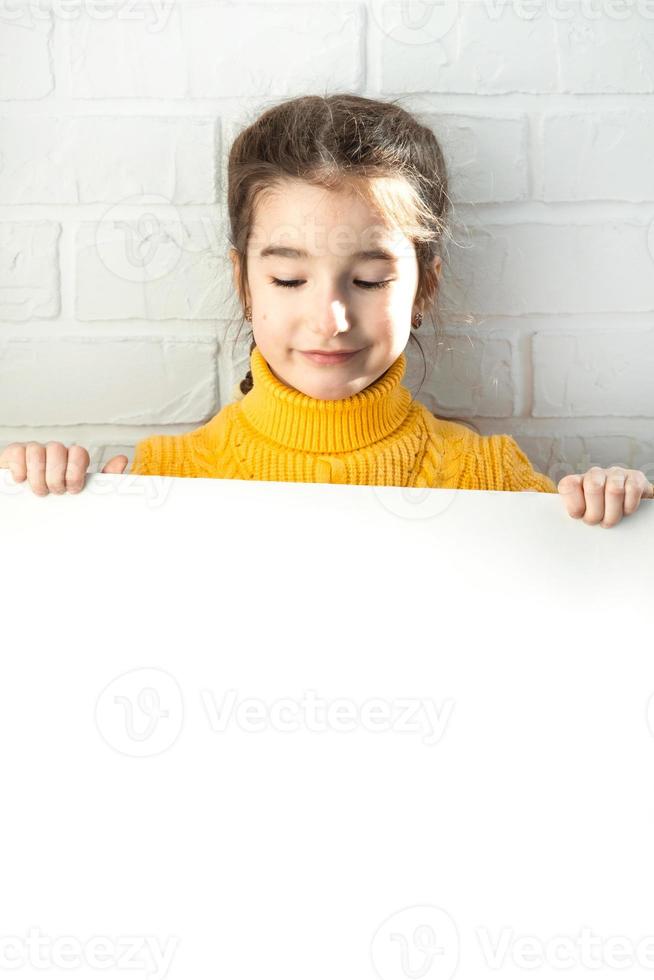 una chica triste sostiene una hoja blanca de papel - maqueta para publicidad, eslogan, inscripción. el espacio de copia está en manos de los niños, el niño está molesto y escondido. foto