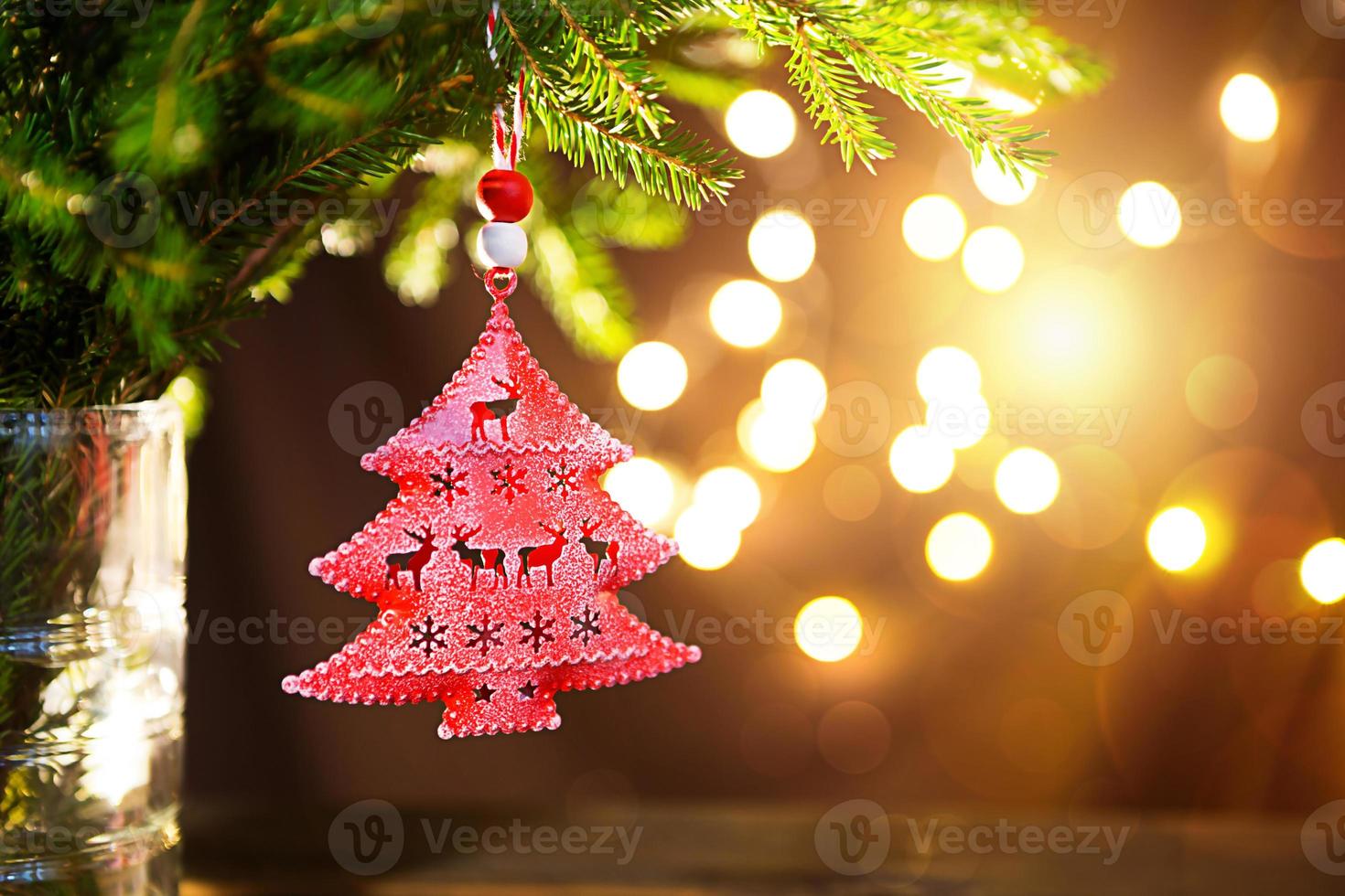 Red Christmas tree toy on a branch of a natural fir tree with lights of garlands in defocus in the background. Metal toy with slits of deer and snowflakes. Christmas, new year, copy space, bokeh. photo