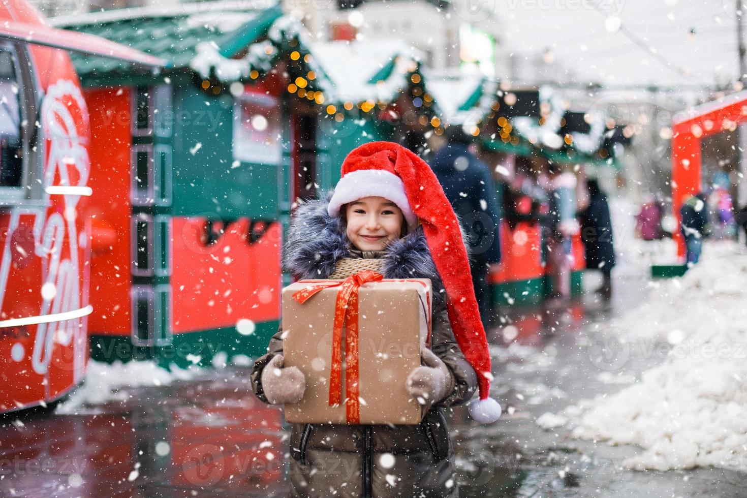 Portrait of joyful girl in Santa hat with gift box for Christmas on city street in winter with snow on festive market with decorations and fairy lights. Warm clothes, knitted scarf and fur. New year photo