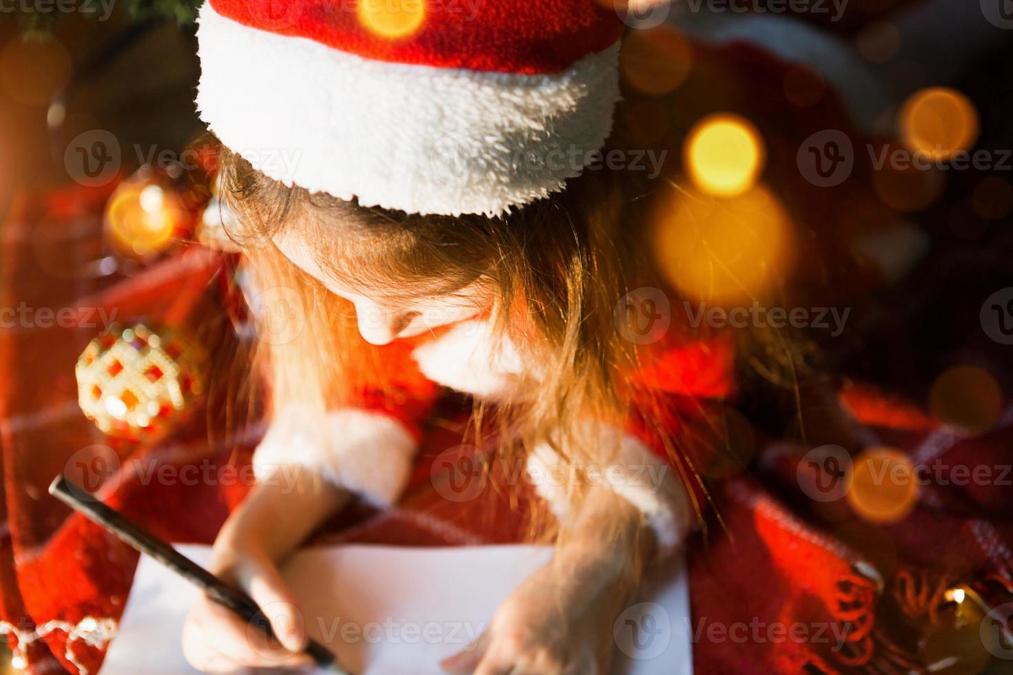 Little girl in a Santa hat and red dress under Christmas tree is dreaming, waiting for the holiday, lying on a plaid blanket. A letter on piece of paper, gifts. New year, Christmas. defocus lights photo