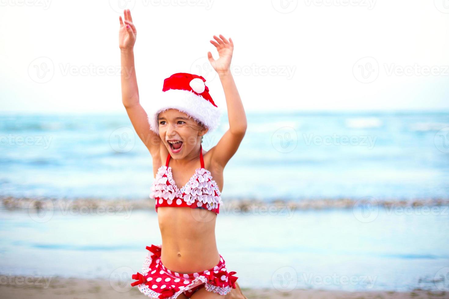 Una niña de 5 años con traje de baño rojo y gorro de Papá Noel en la playa está feliz, grita, salta y agita las manos con felicidad. viajes de invierno de navidad y año nuevo a países cálidos. turismo para vacaciones en trópicos foto