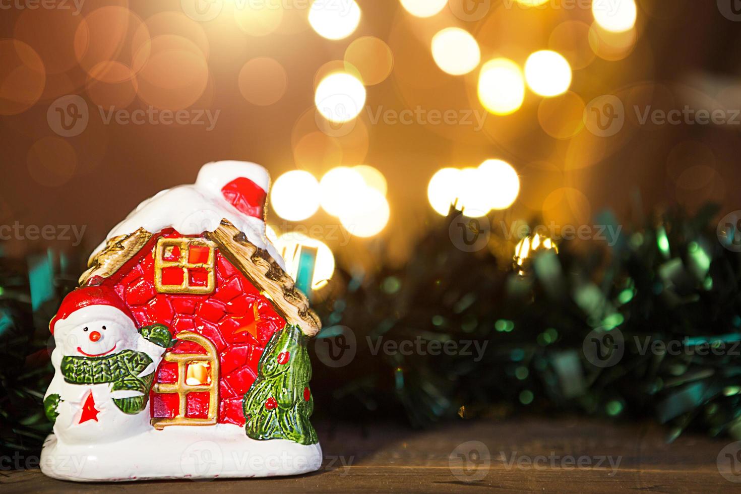Cozy red gingerbread house with a light in the window and a snowman on a wooden background with copyspace and defocus lights in bokeh. New year, Christmas, holiday background, home photo