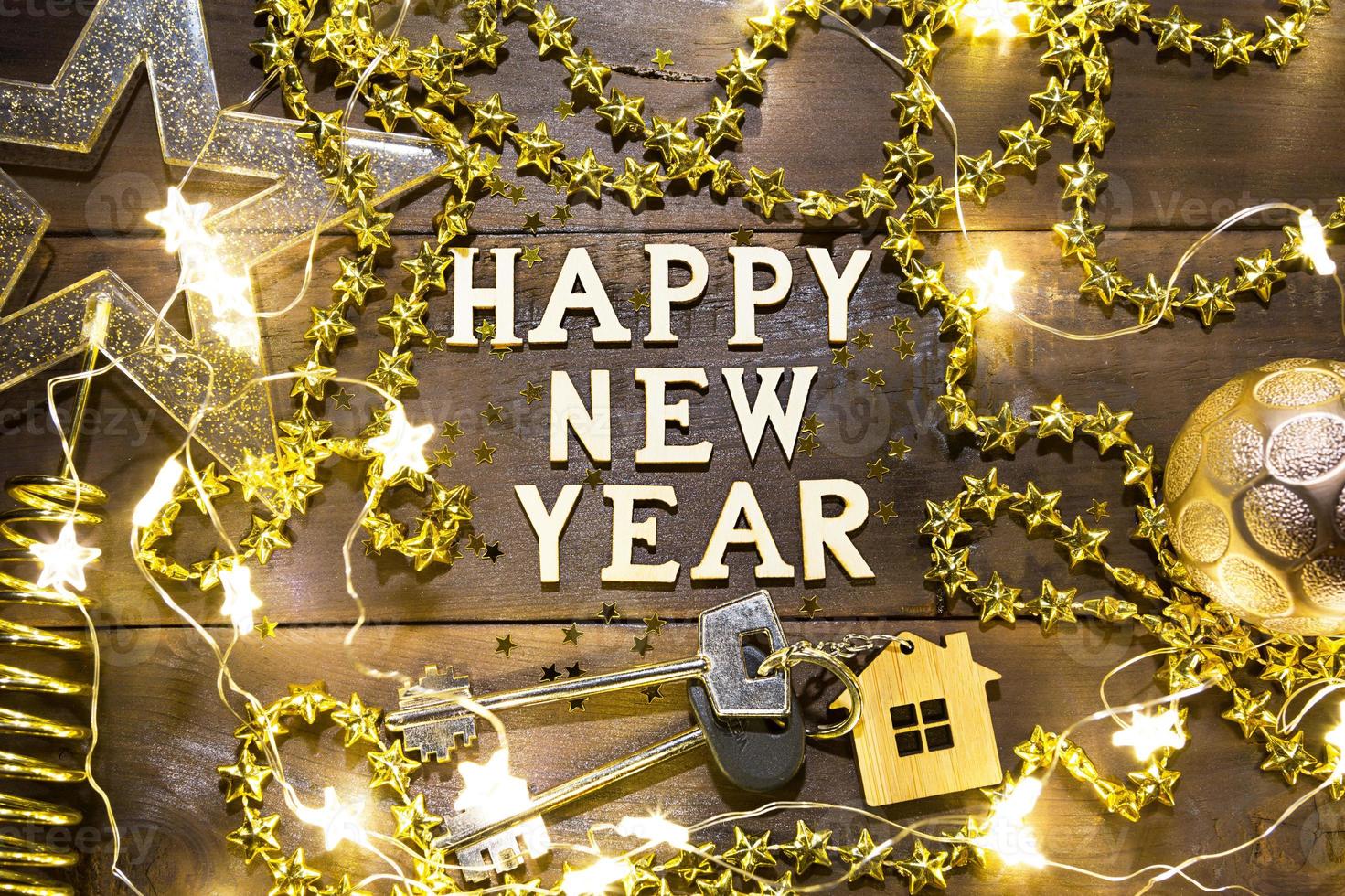 House key with keychain cottage on a festive background with sequins, stars, lights of garlands. Happy New Year-wooden letters, greetings, greeting card. Purchase, construction, relocation, mortgage photo