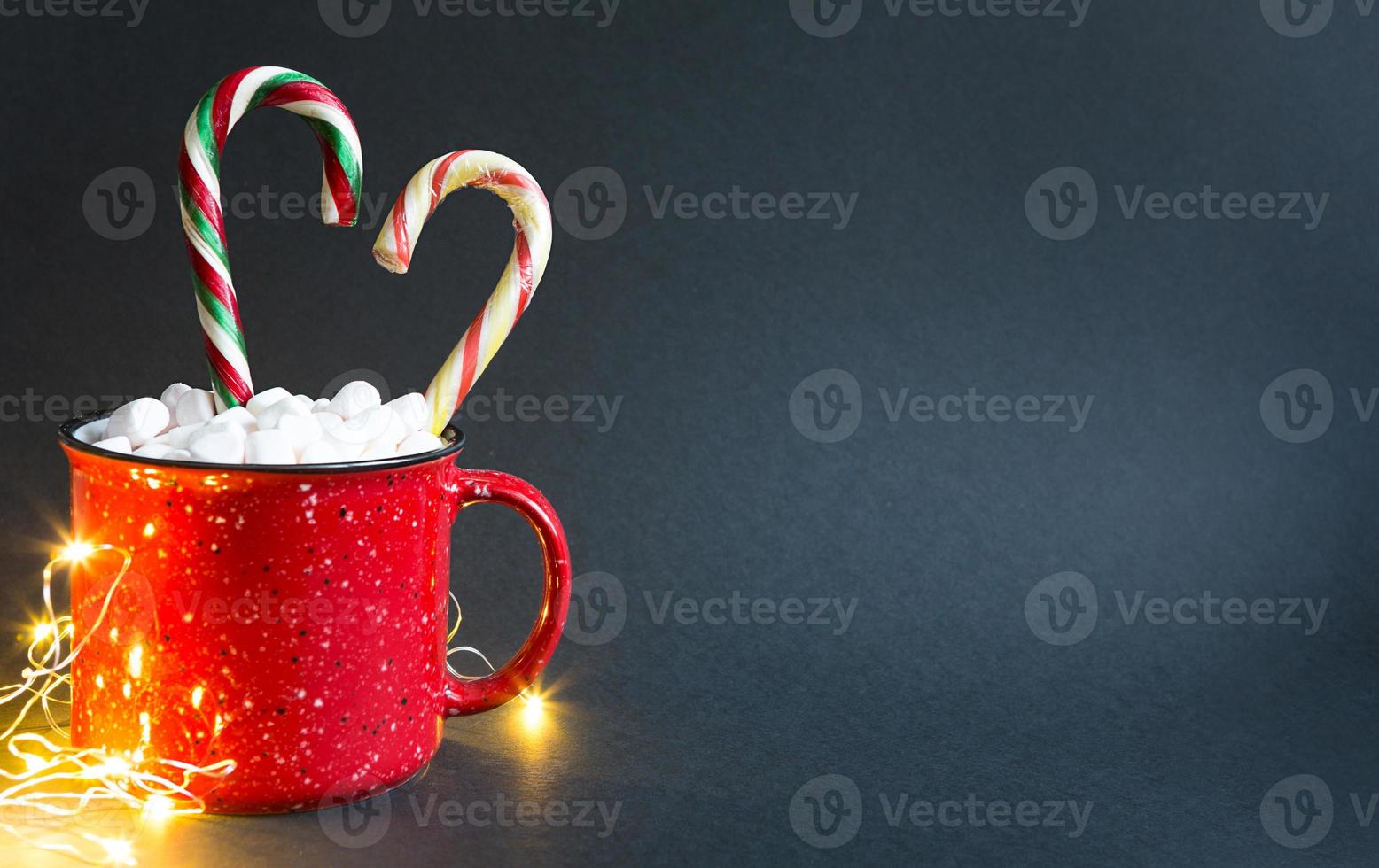 Red mug with marshmallows and caramel candy cane in the shape of a heart and lights of garlands on a black background. Christmas, New Year, festive mood. Copy space photo