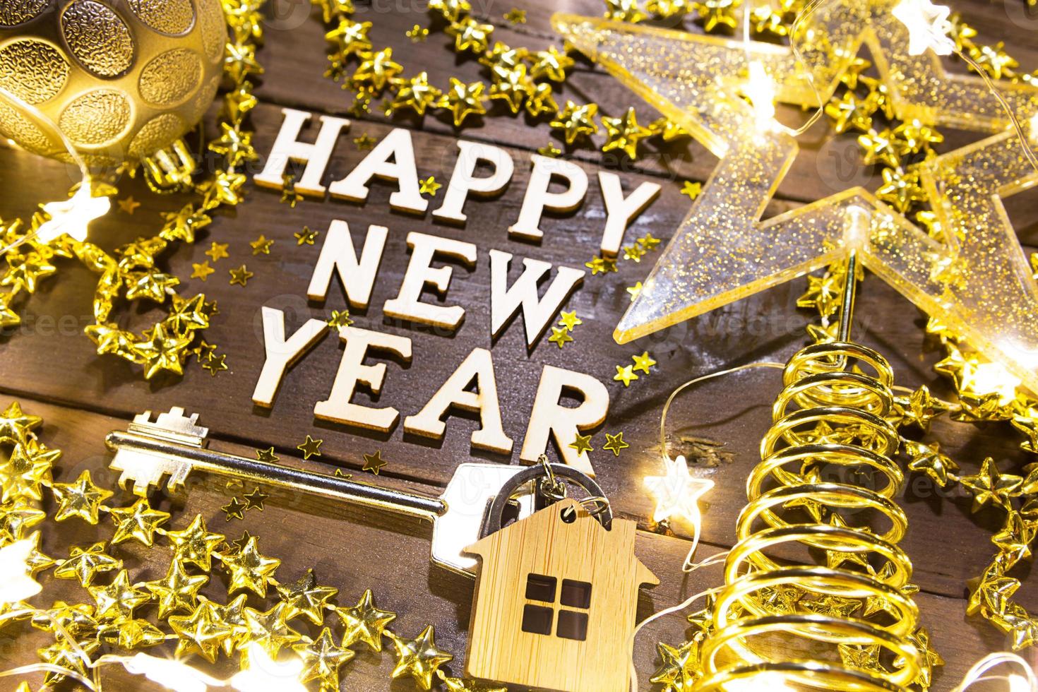 House key with keychain cottage on a festive background with sequins, stars, lights of garlands. Happy New Year-wooden letters, greetings, greeting card. Purchase, construction, relocation, mortgage photo