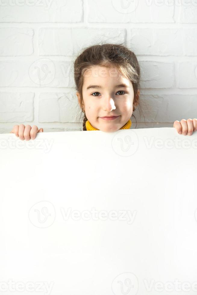 una chica triste sostiene una hoja blanca de papel - maqueta para publicidad, eslogan, inscripción. el espacio de copia está en manos de los niños, el niño está molesto y escondido. foto