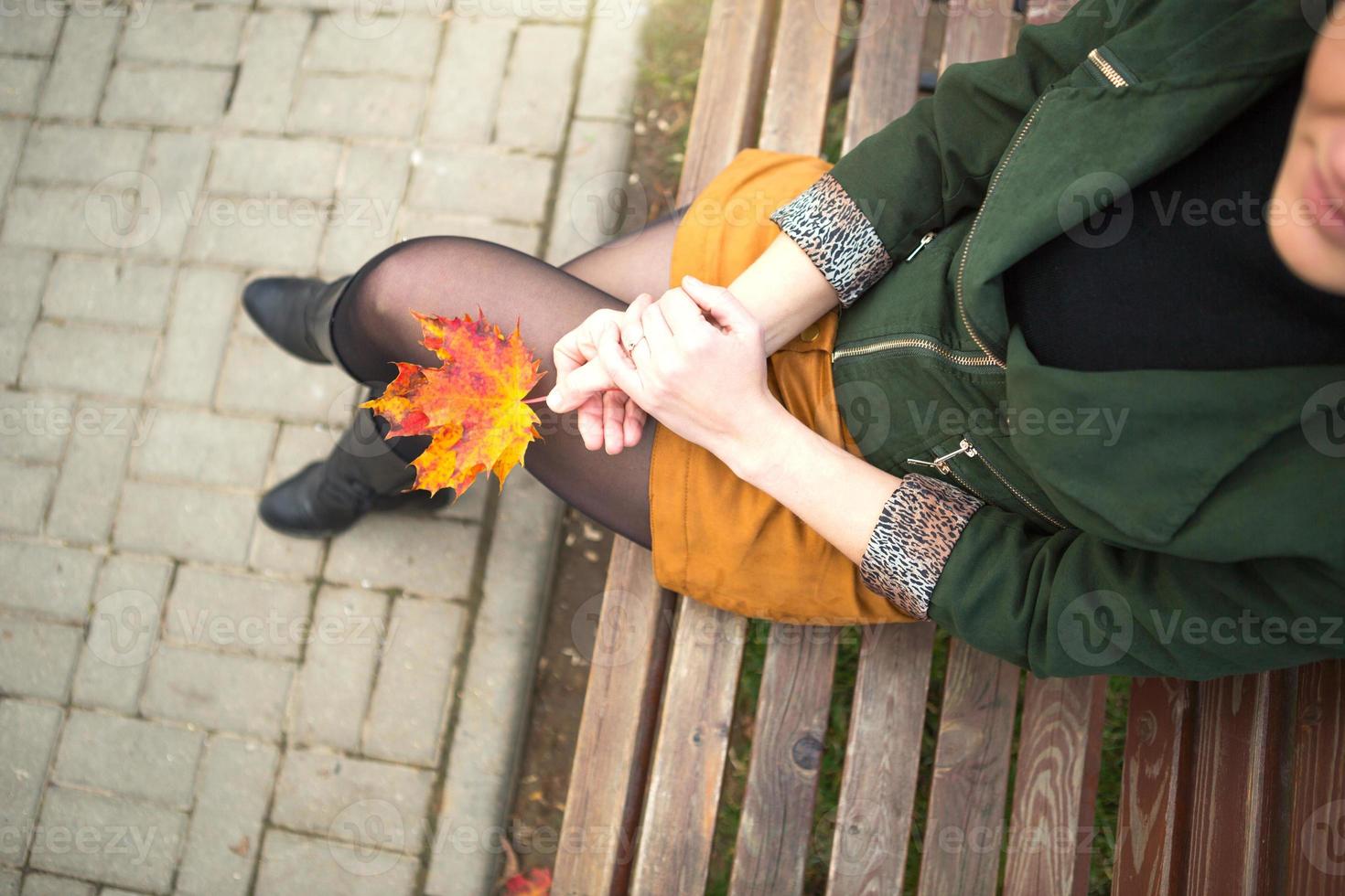 Legs of a woman in nylon tights in a closed pose leg over leg in a short mustard mini skirt and in a green jacket with a maple leaf in her hand. Autumn walk on a park bench. Autumn mood and fashion photo